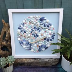 a framed art piece with shells and succulents on it next to a potted plant