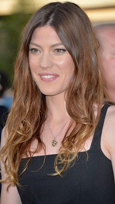 a woman with long brown hair wearing a black dress and smiling at the camera while standing in front of other people