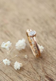 two gold wedding bands with diamonds on them sitting next to white flower petals in front of a wooden surface