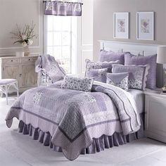 a bed with purple bedspread and pillows in a white room next to a window