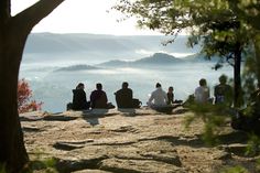 several people sitting on top of a large rock with mountains in the background and fog