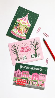 three christmas cards and a pencil on a white surface with holiday greetings written below them