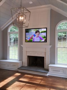 a television mounted on the wall above a fireplace in a room with two windows and a chandelier