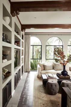 a living room filled with lots of furniture next to large window covered in plants and bookshelves