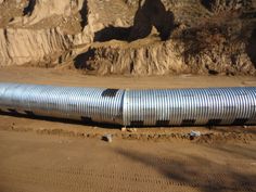 a large metal pipe laying on top of a dirt field next to a rocky cliff