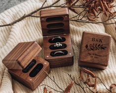 three wooden boxes with wedding rings in them on a white blanket next to dried leaves