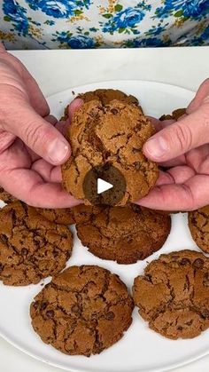 a person holding a chocolate chip cookie in front of several cookies on a white plate