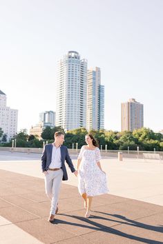 a man and woman holding hands while walking in front of tall buildings on a sunny day