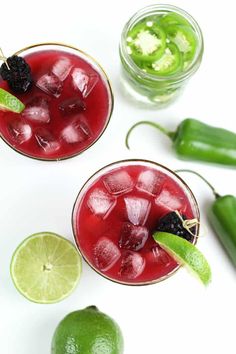 two glasses filled with red and green alcoholic drinks next to limes, jalapenos, and cucumbers