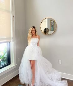 a woman in a white dress standing next to a window with a mirror on the wall