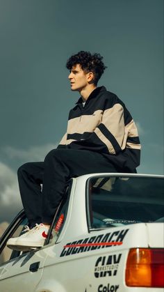 a man sitting on top of a white car in front of a cloudy blue sky