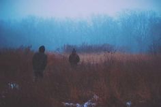 two people standing in the middle of a field on a foggy day with trees behind them