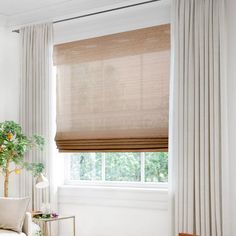 a living room with white curtains and a window covered in beige drapes, next to a plant