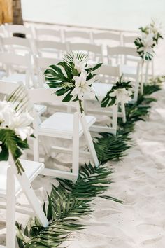 the wedding aisle is decorated with white chairs and greenery for an elegant beach ceremony