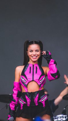 a woman in pink and black outfit standing next to a drum set with one hand on her hip