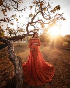 a woman in an orange dress is standing near a tree with the sun behind her