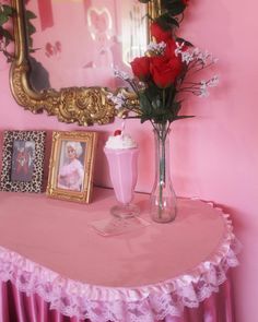 a pink table topped with a vase filled with flowers next to a mirror and framed pictures