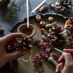 two hands are decorating pine cones on a tray with berries and other holiday decorations