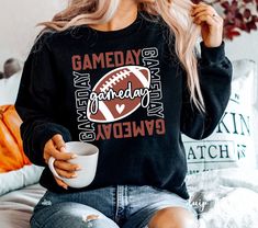 a woman sitting on a couch holding a coffee cup and wearing a gameday sweatshirt