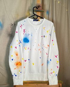 a white sweatshirt with paint splattered all over it hanging on a clothes rack