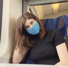 a woman wearing a blue face mask sitting on a train with her hands behind her head