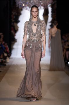 a woman walking down a runway wearing a dress with chains on it's shoulders