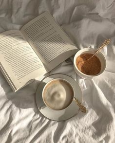 an open book and two cups of coffee sit on a bed next to each other