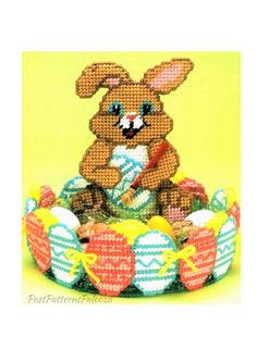 a cross stitch bunny sitting on top of an easter basket with eggs and carrots