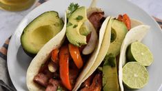 three tacos on a plate with sliced avocado, peppers and cilantro