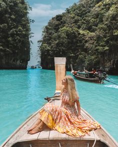 a woman sitting on top of a wooden boat in the middle of blue water with trees