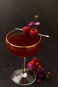 two raspberries sit on top of a drink in a coupe glass with some garnish