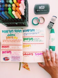 a person's hand holding an open planner next to markers, pens and markers