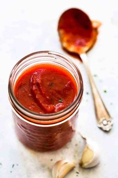 a jar filled with red sauce next to garlic on a white tablecloth and two spoons