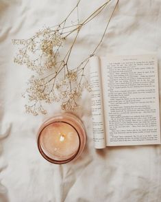 an open book next to a candle and some flowers on a white sheeted surface