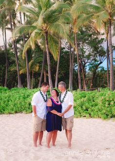 three people standing on the beach with palm trees in the background and one person wearing a lei