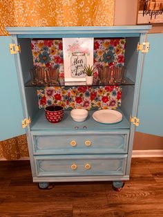 a blue dresser with flowers on the wall and dishes in it's cupboards