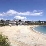 a sandy beach with houses on the shore and blue water in the foreground, under a partly cloudy sky