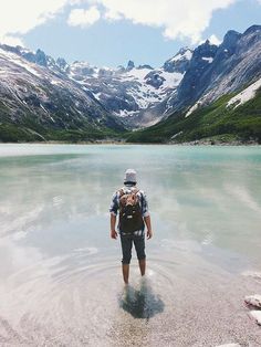 a man with a backpack standing in the middle of a lake surrounded by snow capped mountains