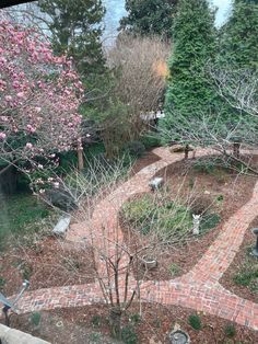 a brick path in the middle of a garden surrounded by trees and shrubs with pink flowers