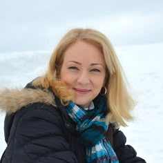 a woman with blonde hair wearing a black jacket and blue scarf standing in the snow