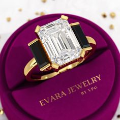 an emerald cut diamond ring sits on top of a purple velvet box with gold and black accents