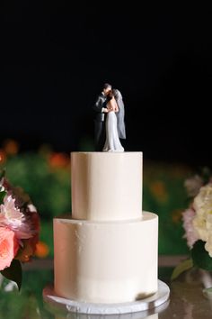 a bride and groom figurine on top of a wedding cake with flowers in the background