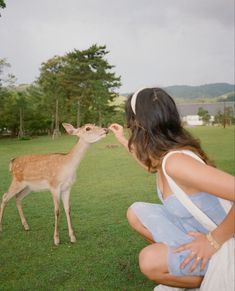 a woman kneeling down next to a deer on top of a grass covered field with trees in the background