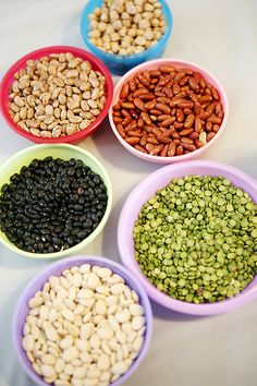 seven bowls filled with different types of beans and nuts on a table top, all in various colors