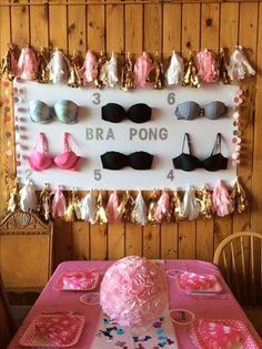 a pink table topped with lots of cake next to a wall mounted bra pong sign