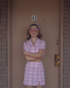 a woman standing in front of a door with her arms crossed, wearing sunglasses and a pink dress