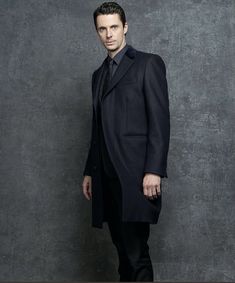 a man in a black suit and tie standing against a wall with his hands in his pockets