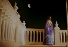 a woman standing on a balcony at night with the moon in the sky behind her