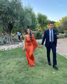 a man and woman in formal wear holding hands while walking through the grass with trees behind them