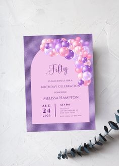 a pink and purple birthday party card with balloons on the front, sitting next to some flowers